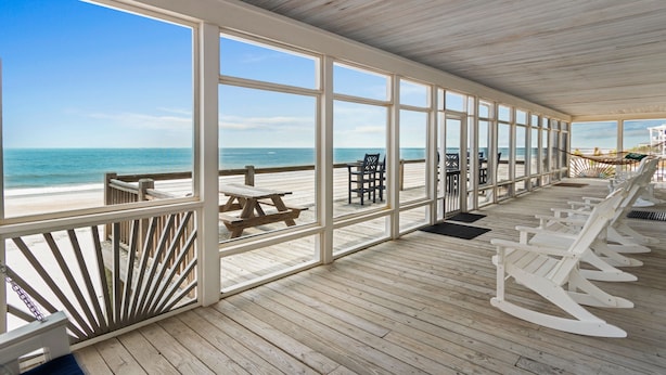 A covered deck with wooden flooring, large windows, and white rocking chairs facing the ocean. - 6-Bedroom Beach House w/ Private Dock & Pool in Inlet Harbour (Murrells Inlet)