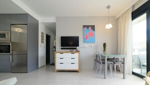 A modern living room with a flat-screen TV, a dining area with clear chairs, and a kitchen with a built-in oven and microwave.