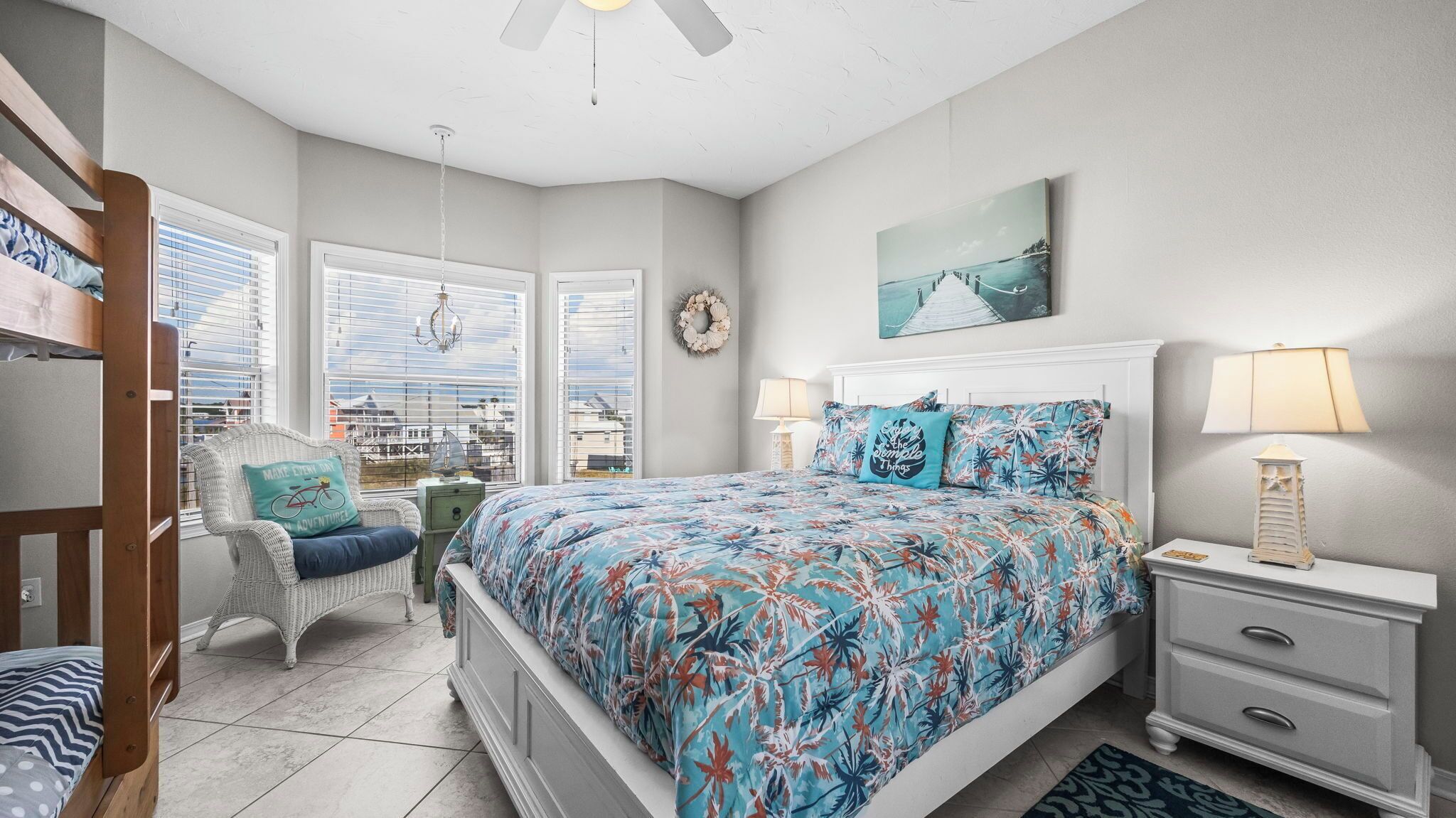 A bedroom with a bed, two bedside tables, a chair, a window with blinds, and a view of the city.