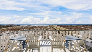 Une terrasse au bord de la plage, avec des chaises et des tables Adirondack blanches, donnant sur une plage de sable et une promenade en bois menant à l’océan.