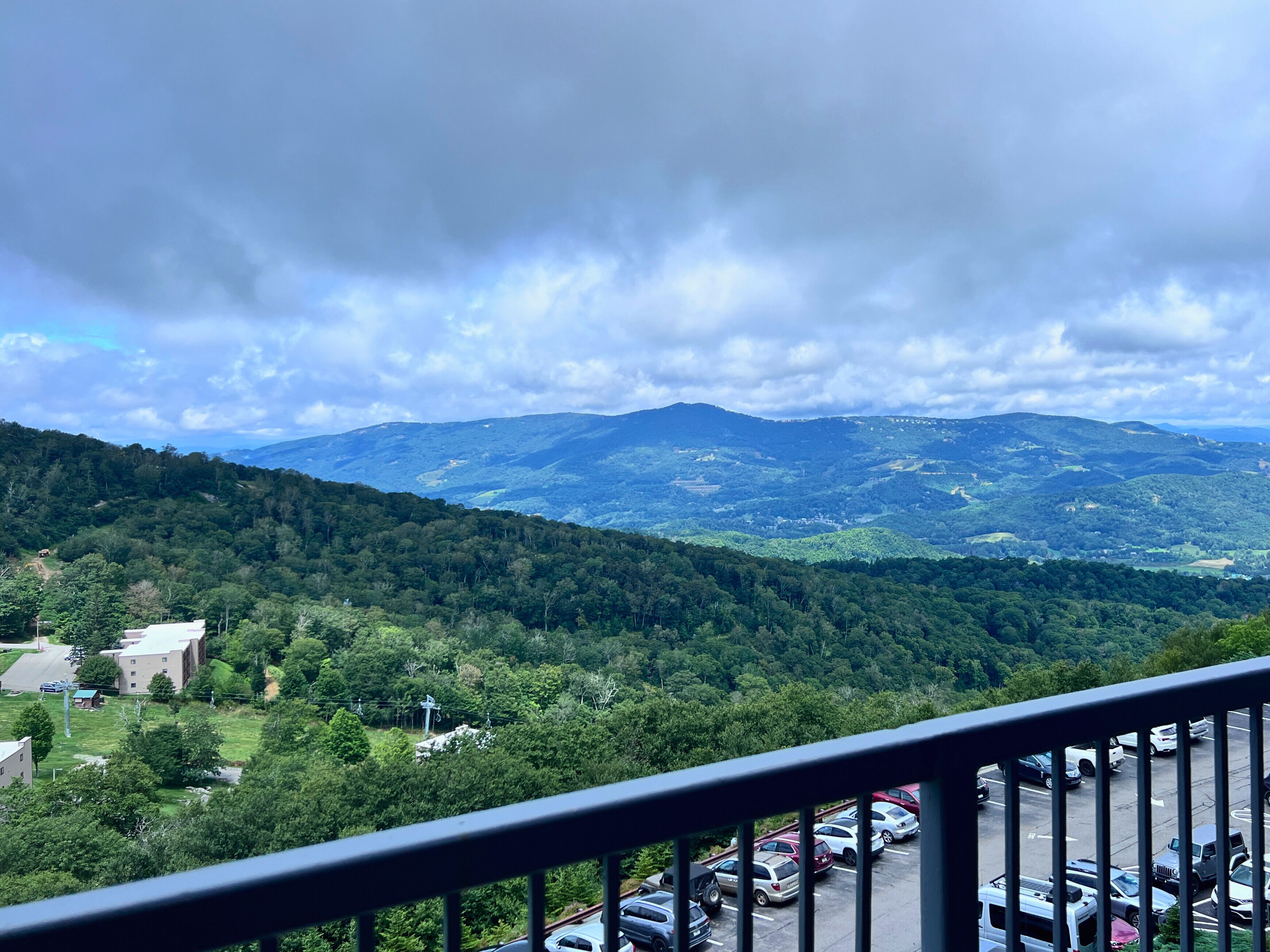 A view from a balcony overlooking a parking lot and a mountainous landscape with dense forests.