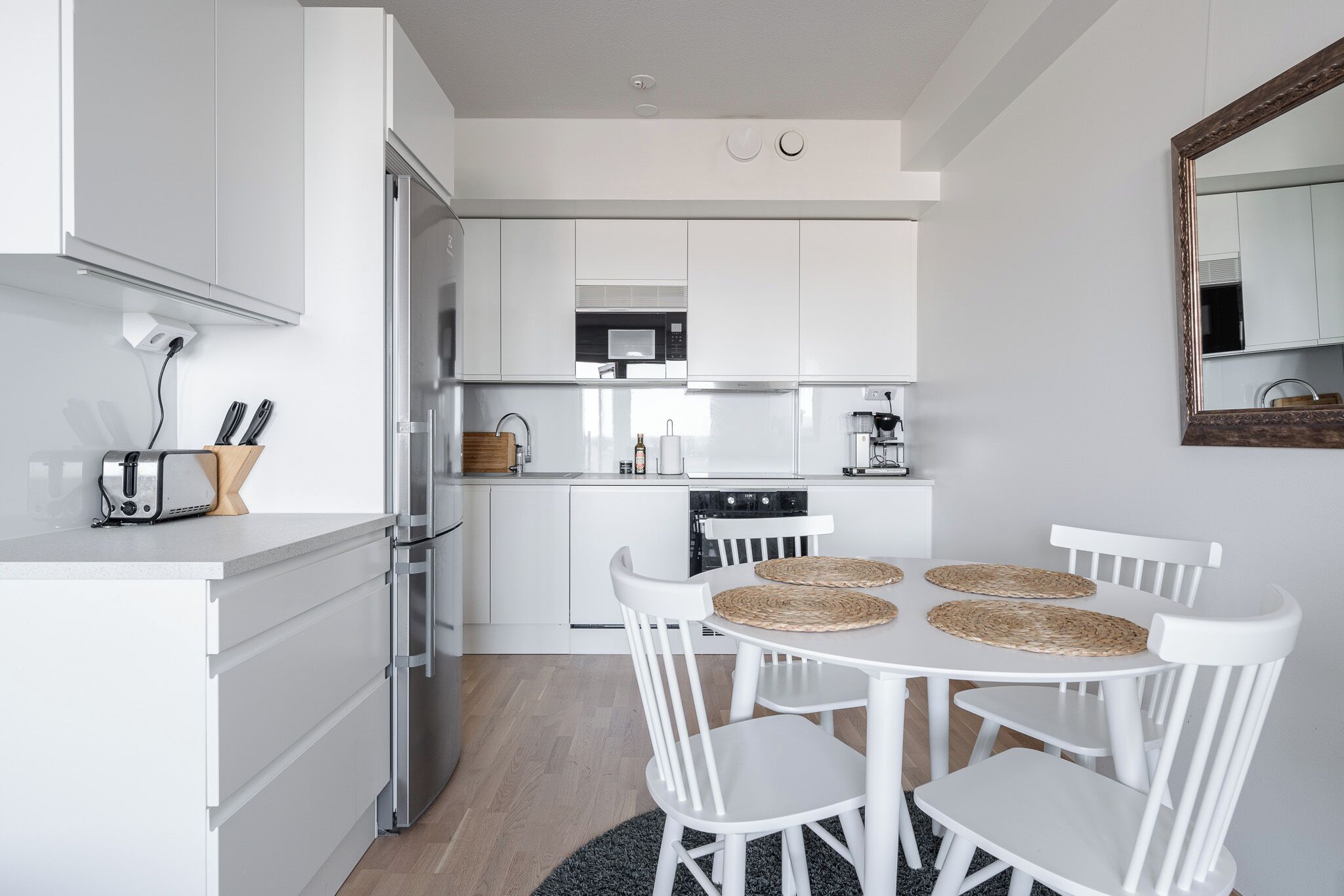 A modern kitchen with white cabinetry, a dining table with four chairs, and a large mirror on the wall.