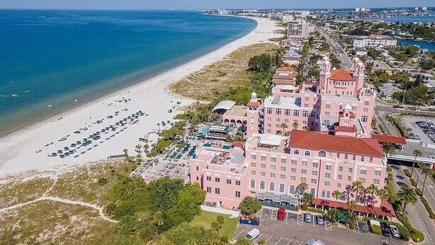 Vista aérea de un hotel frente al mar, un edificio grande con techo rojo y un estacionamiento.