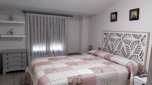 Une chambre à coucher comprenant un lit, une table de chevet, une commode, une fenêtre avec des rideaux et un mur orné de deux tableaux encadrés.