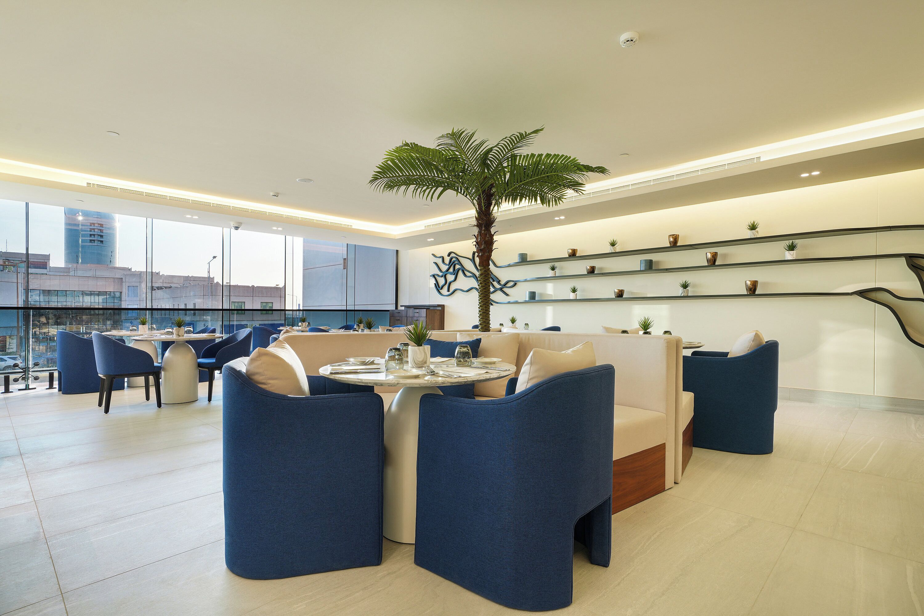 A modern lounge area with a round table, blue chairs, a palm tree, and a glass railing with plants.