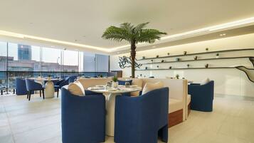 A modern lounge area with a round table, blue chairs, a palm tree, and a glass railing with plants.