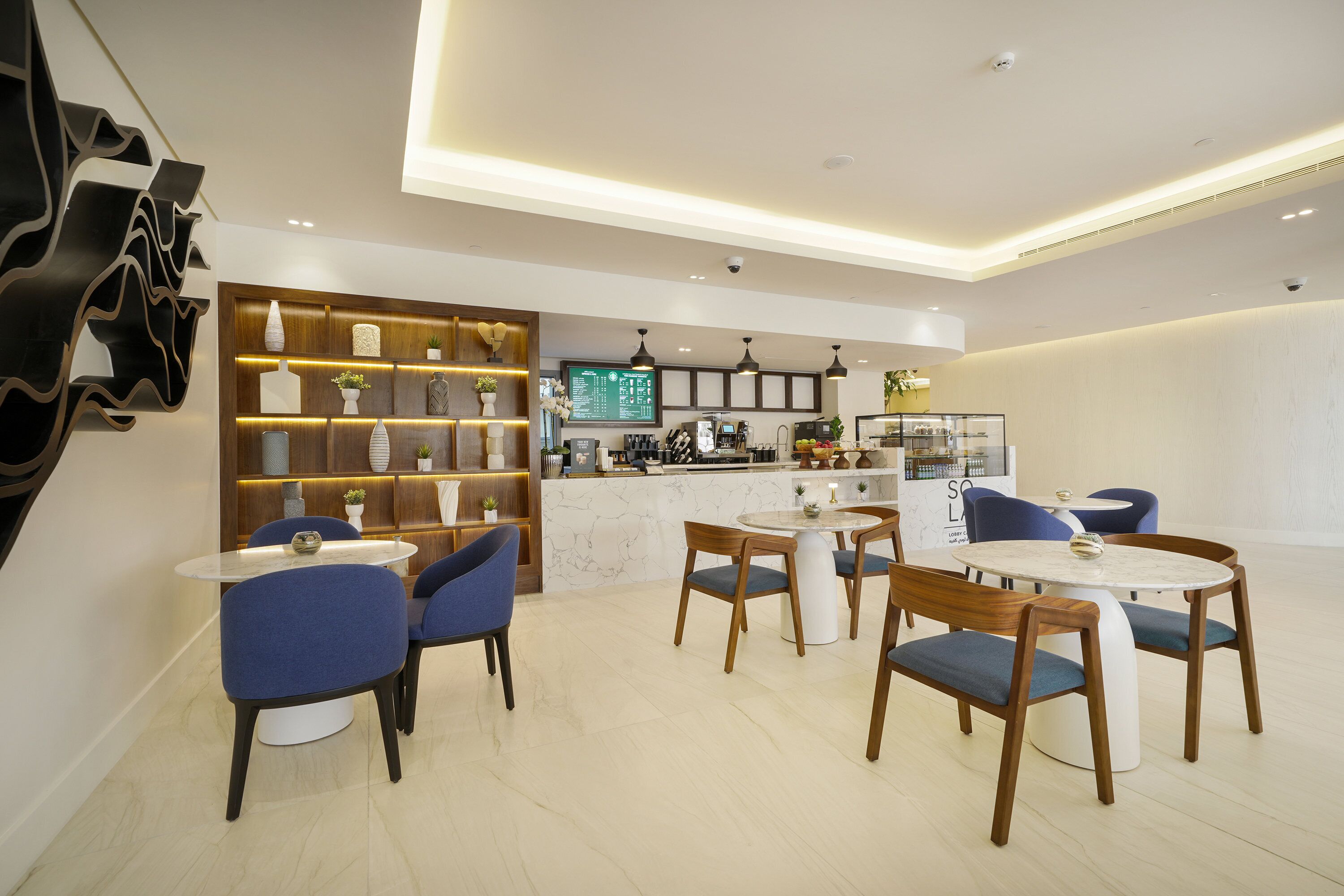 A modern interior with a dining area featuring round tables and blue chairs, a kitchen area with a marble countertop, and a decorative wall with shelves.