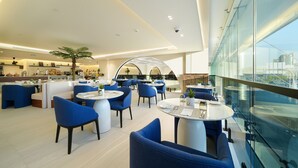 Un hall d’hôtel moderne avec des tables rondes et des chaises bleues, un grand mur de verre et une vue sur la ville.