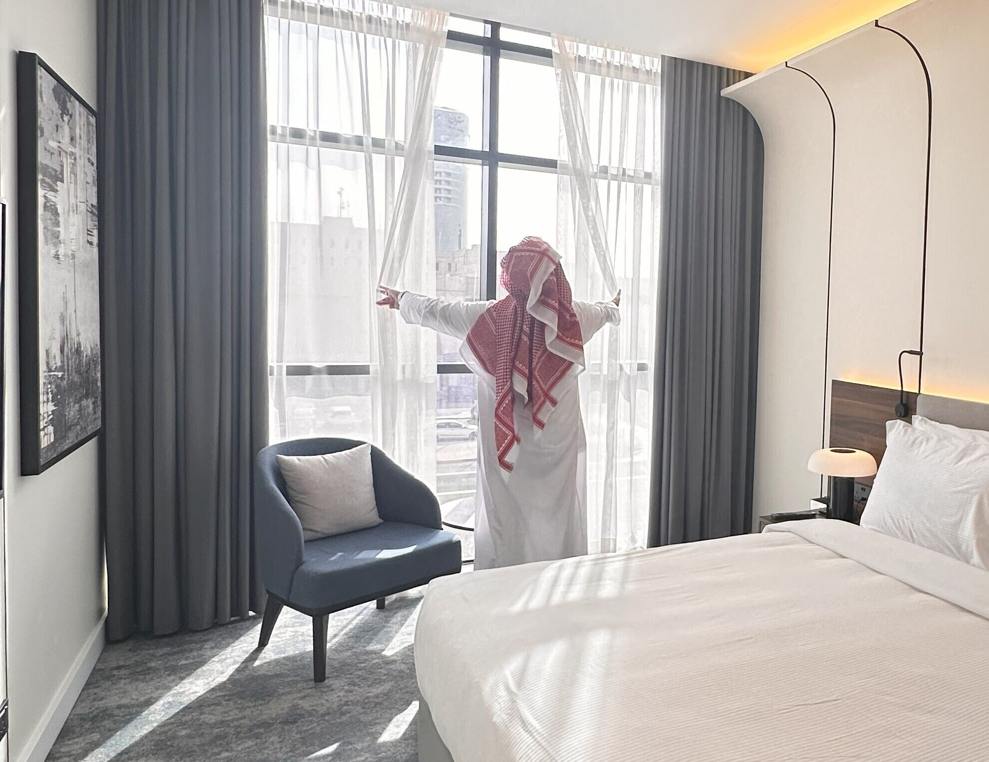 A hotel room with a bed, a chair, a desk with a lamp, and a window with sheer curtains.