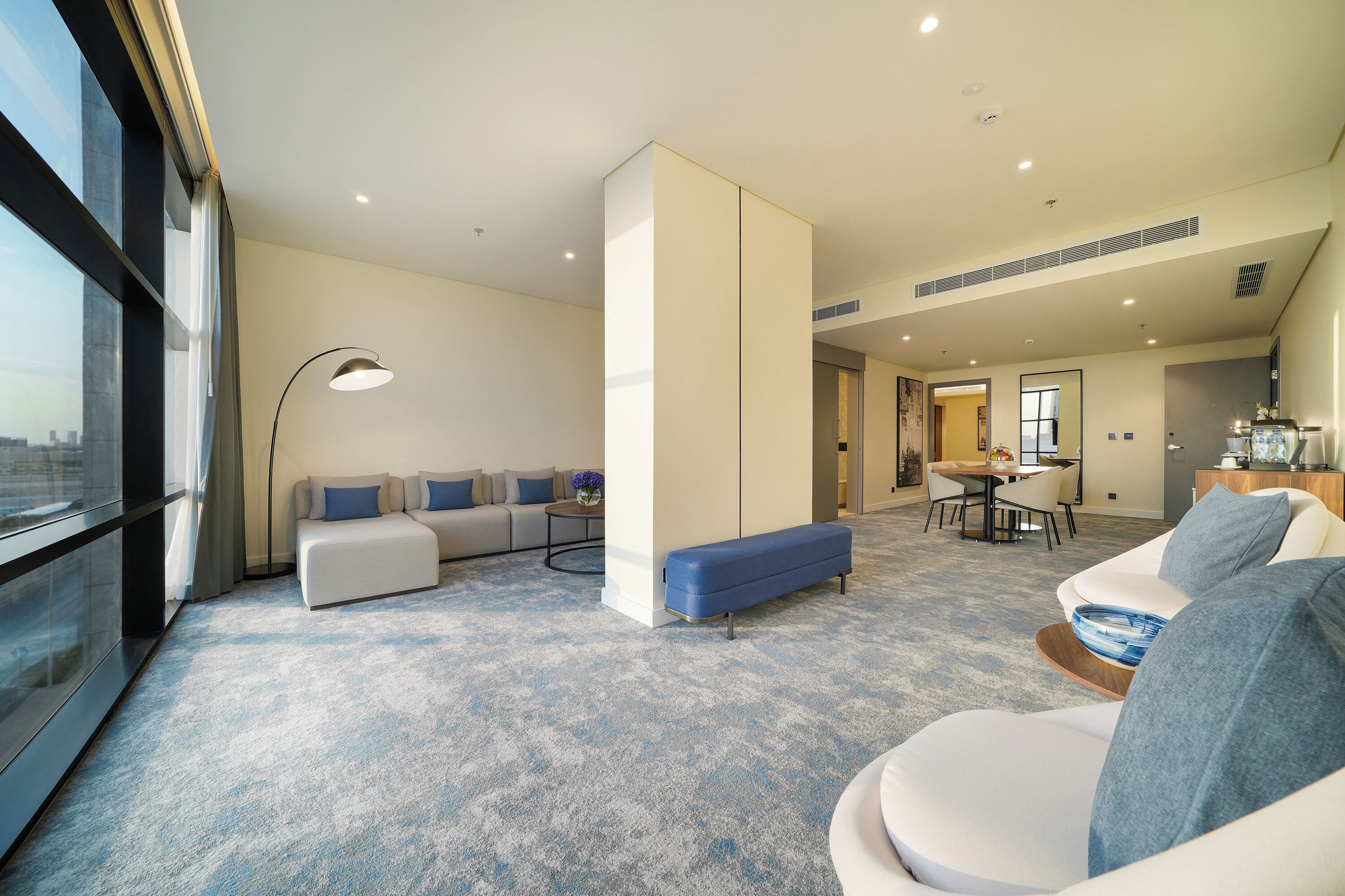 A modern hotel room with a large window, a sofa set, a dining area, and a kitchenette.