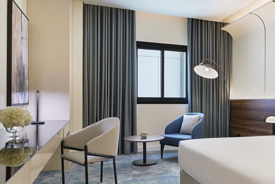 A hotel room with a bed, two chairs, a small table, a lamp, and a window with curtains.