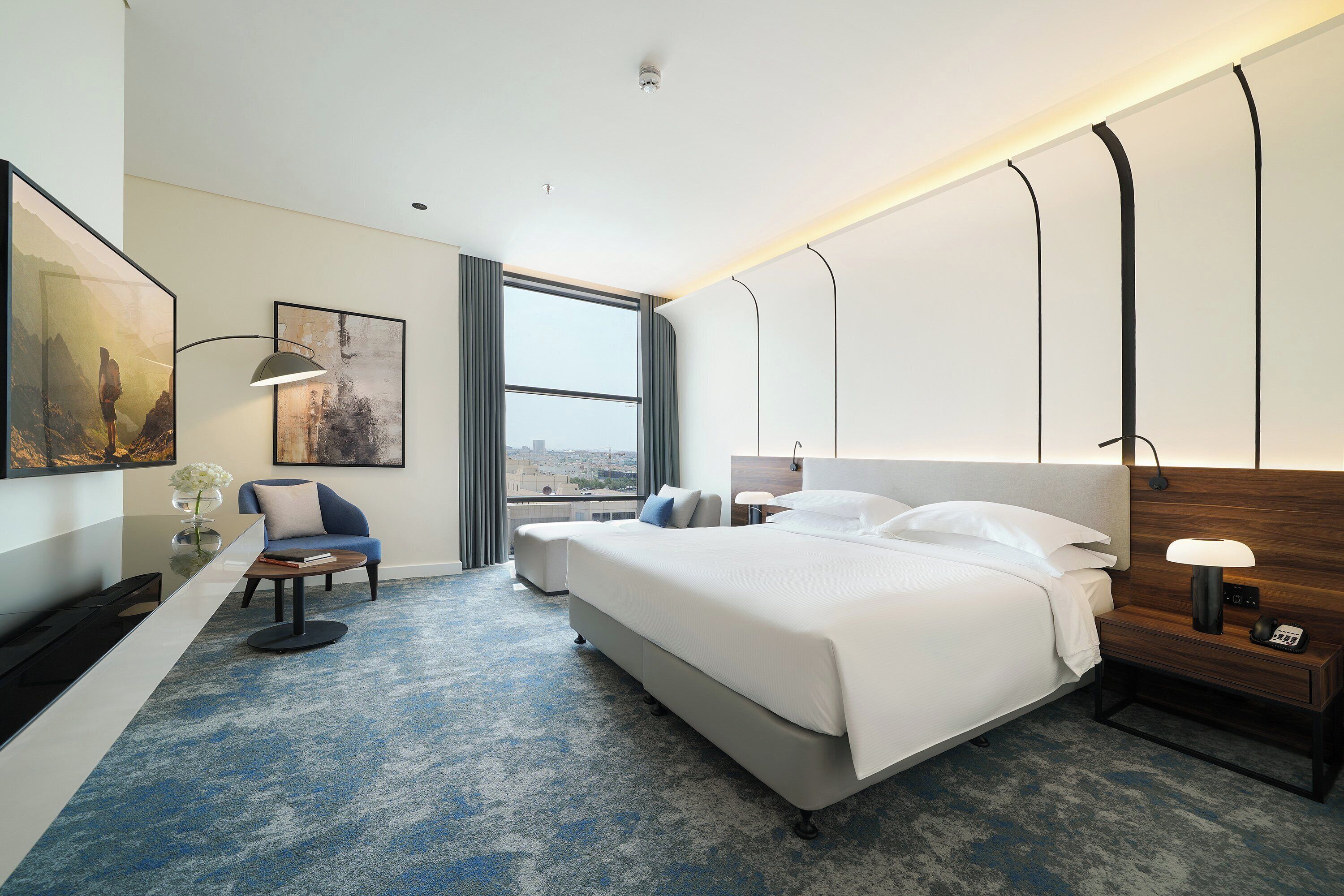 A modern hotel room with a large bed, a desk, a chair, and a view of the city through the window.