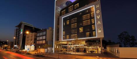 A modern hotel with a prominent 'NOVOM' sign, large windows, and a well-lit entrance area.