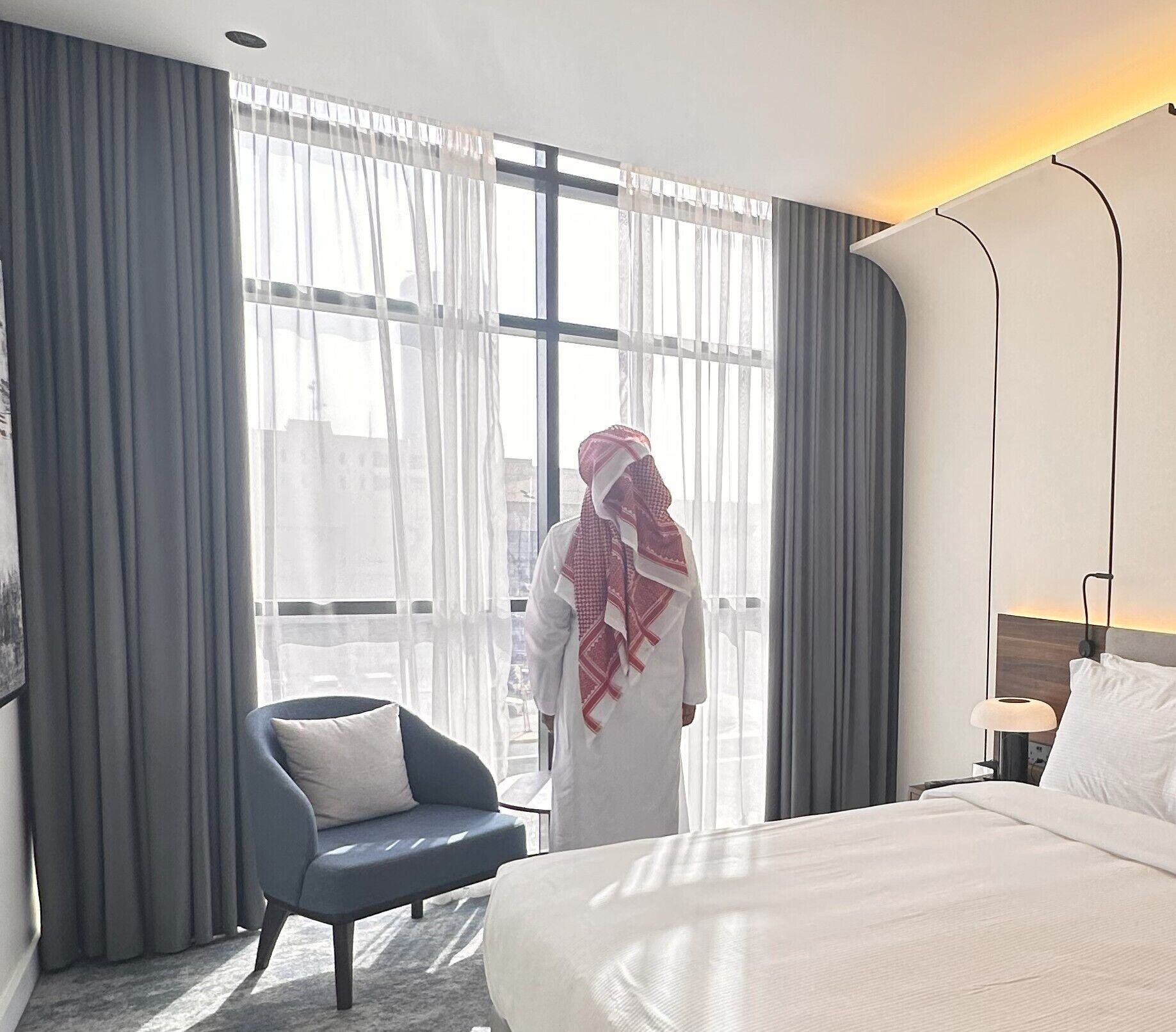 Ett hotellrum med en säng, en stol och ett stort fönster med gardiner.