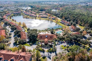 An aerial view of a residential area with a central lake, surrounded by houses with red roofs and palm trees.