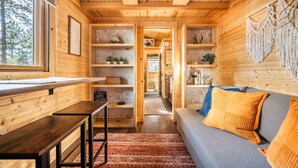 Een gezellig chaletinterieur met houten wanden, een bank met oranje kussens, een eettafel en planken met decoratieve objecten.