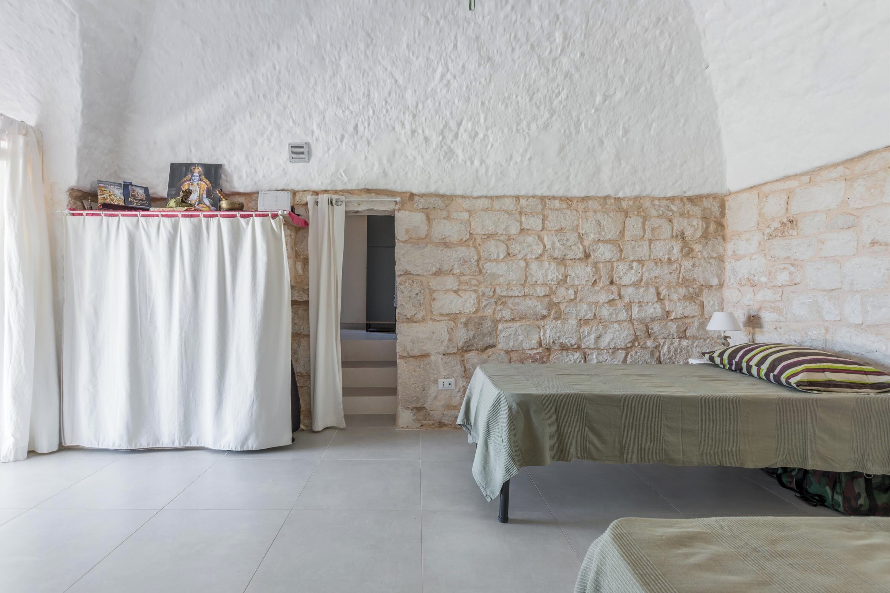 Une pièce avec un mur en pierre, un lit, une petite table avec une lampe et un rideau.