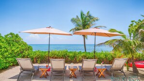 Een terras aan het strand met vier loungesessels en twee parasols, met uitzicht op de oceaan.
