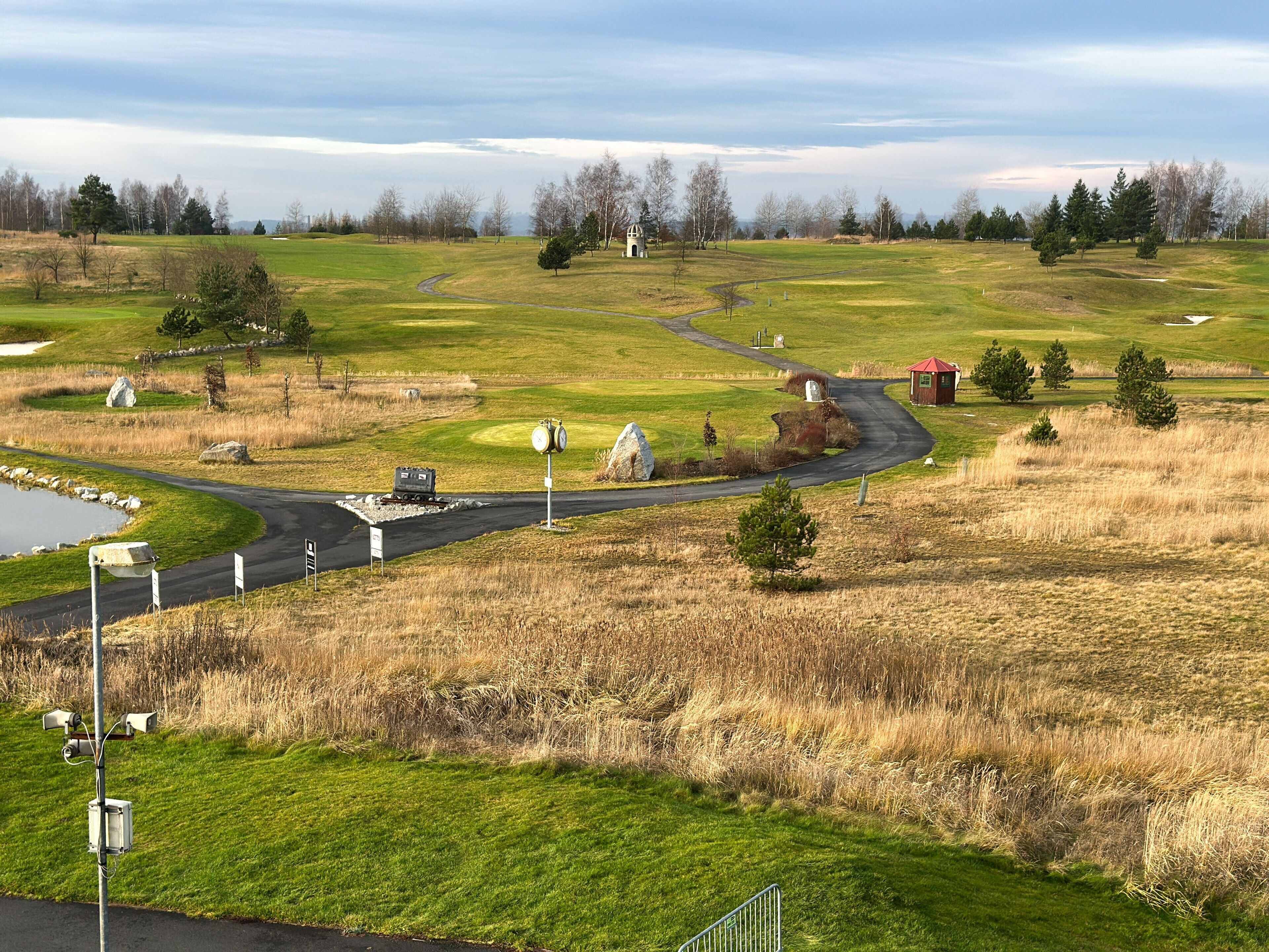 A golf course with a winding road, a pond, and a small red building.