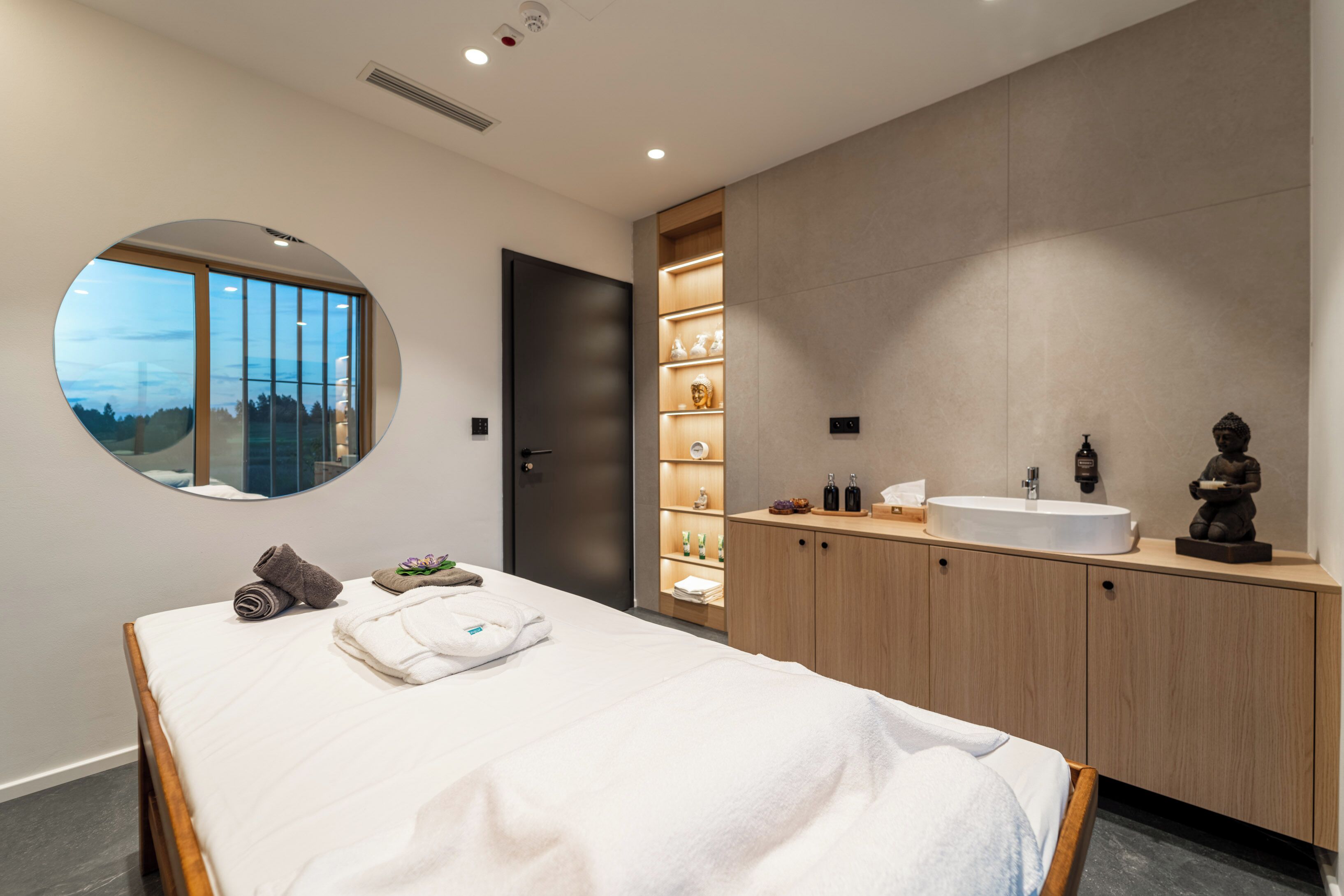 Een moderne slaapkamer met een groot bed, een ronde spiegel, een ingebouwde wastafel en uitzicht op de omgeving door een raam.