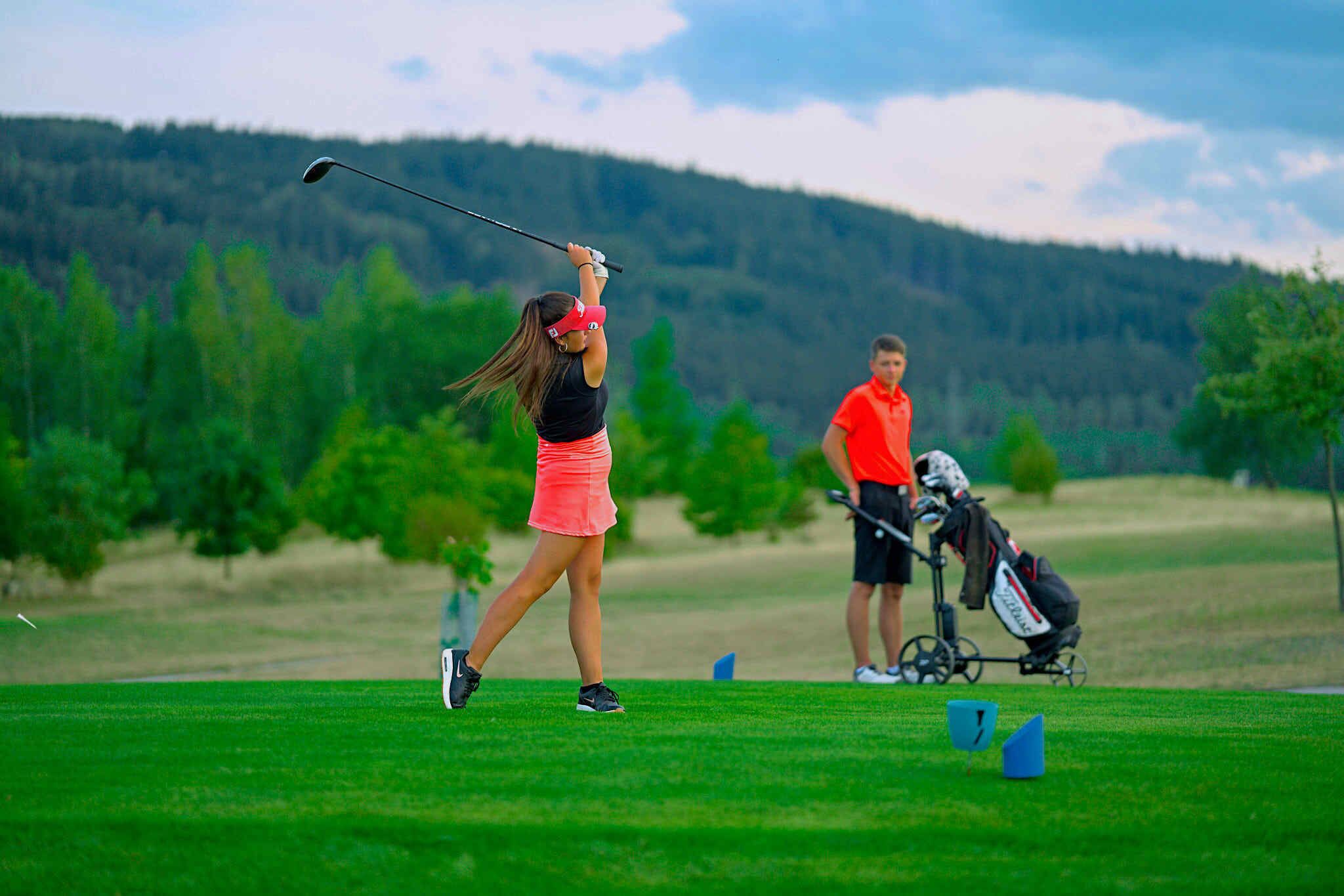 A golf course with a player in mid-swing, a golf bag, and a caddie in the background.
