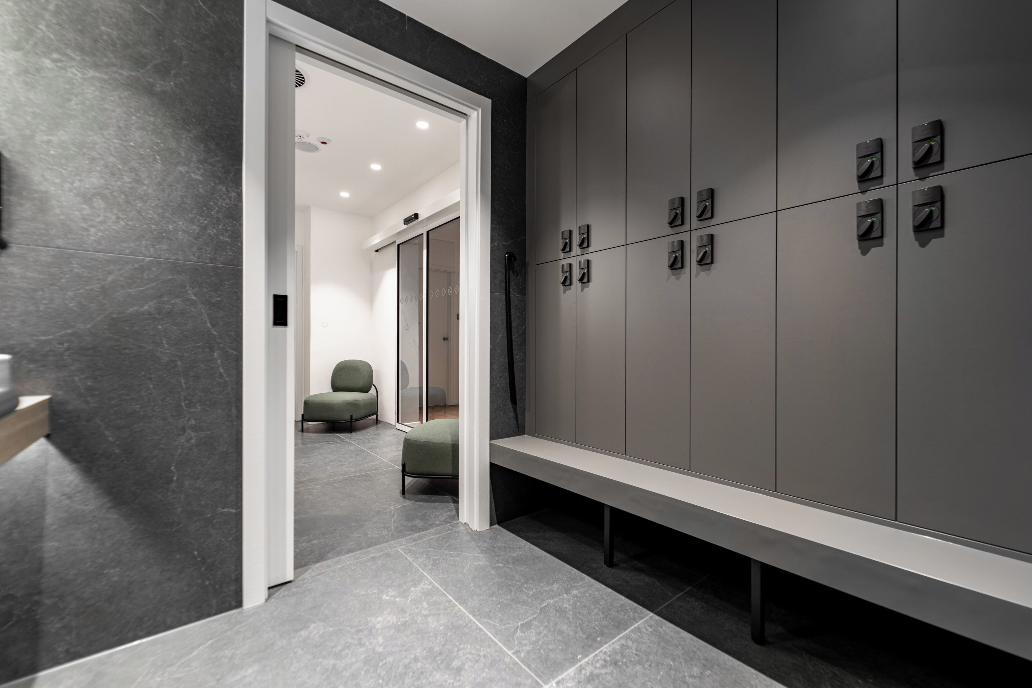 A modern bathroom with grey tiled walls and floor, a bench, and a green chair.