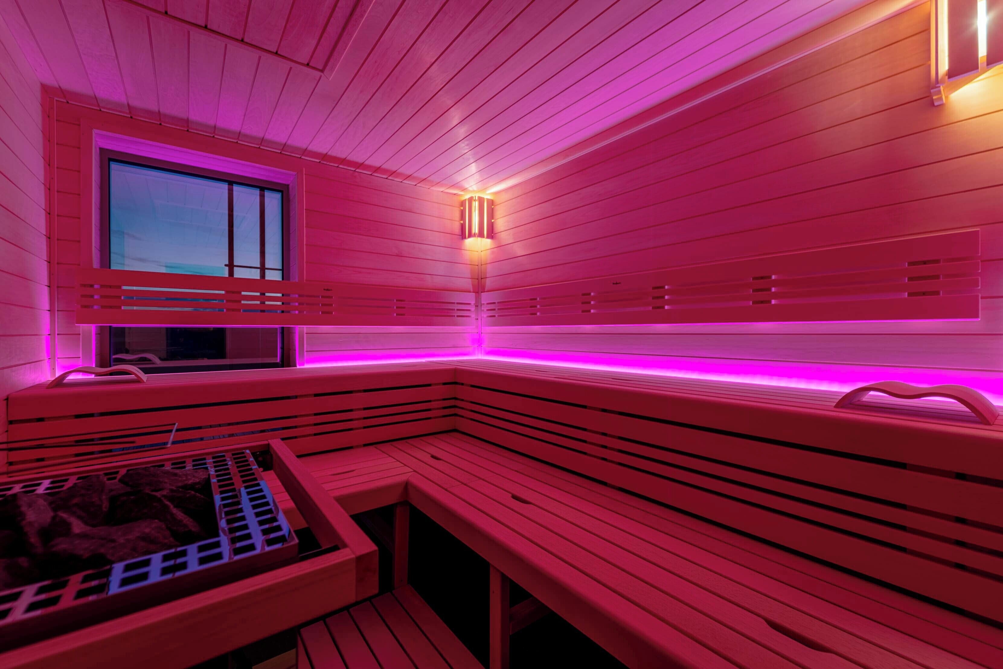 A sauna with wooden benches and a window, illuminated by pink lighting.