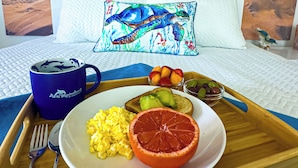 Habitación de hotel con vista al mar, una cama con una almohada con silueta de tortuga marina, una bandeja de desayuno con tostadas, aguacate, huevos y pomelo.