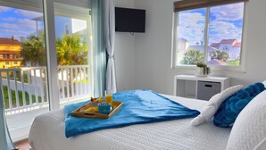 Habitación de hotel con cama, mesita de noche, televisor y balcón con vista a edificios y palmeras.