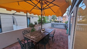 Patio esterno con tavolo e sedie di legno, un ombrello arancione e piante in vaso.