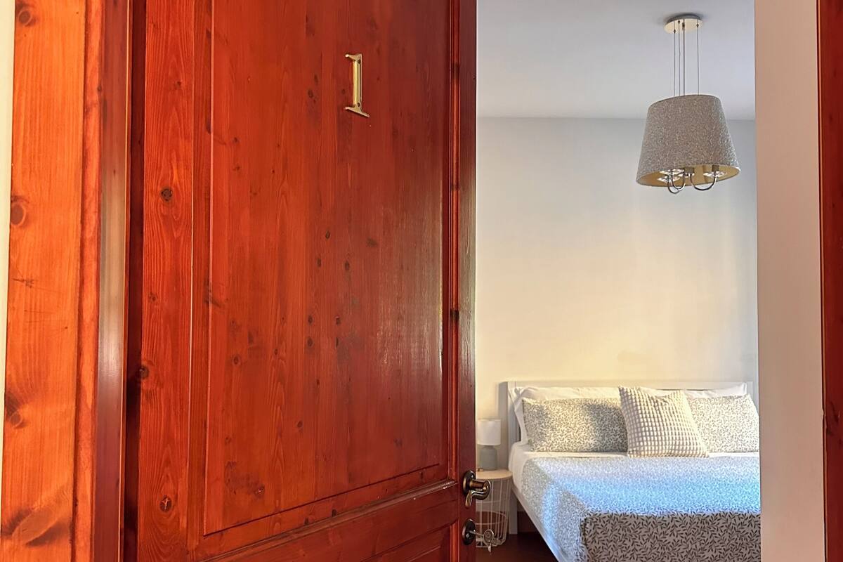 A wooden door with a brass handle and a number plate, leading to a bedroom with a bed, patterned pillows, a bedside table, and a hanging light fixture.
