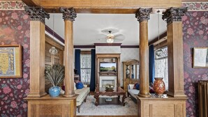 A room with ornate wooden columns, a fireplace, and floral wallpaper. - Grand Historic Retreat | Walk to Capitol & Attractions | Timeless Elegance (Springfield)