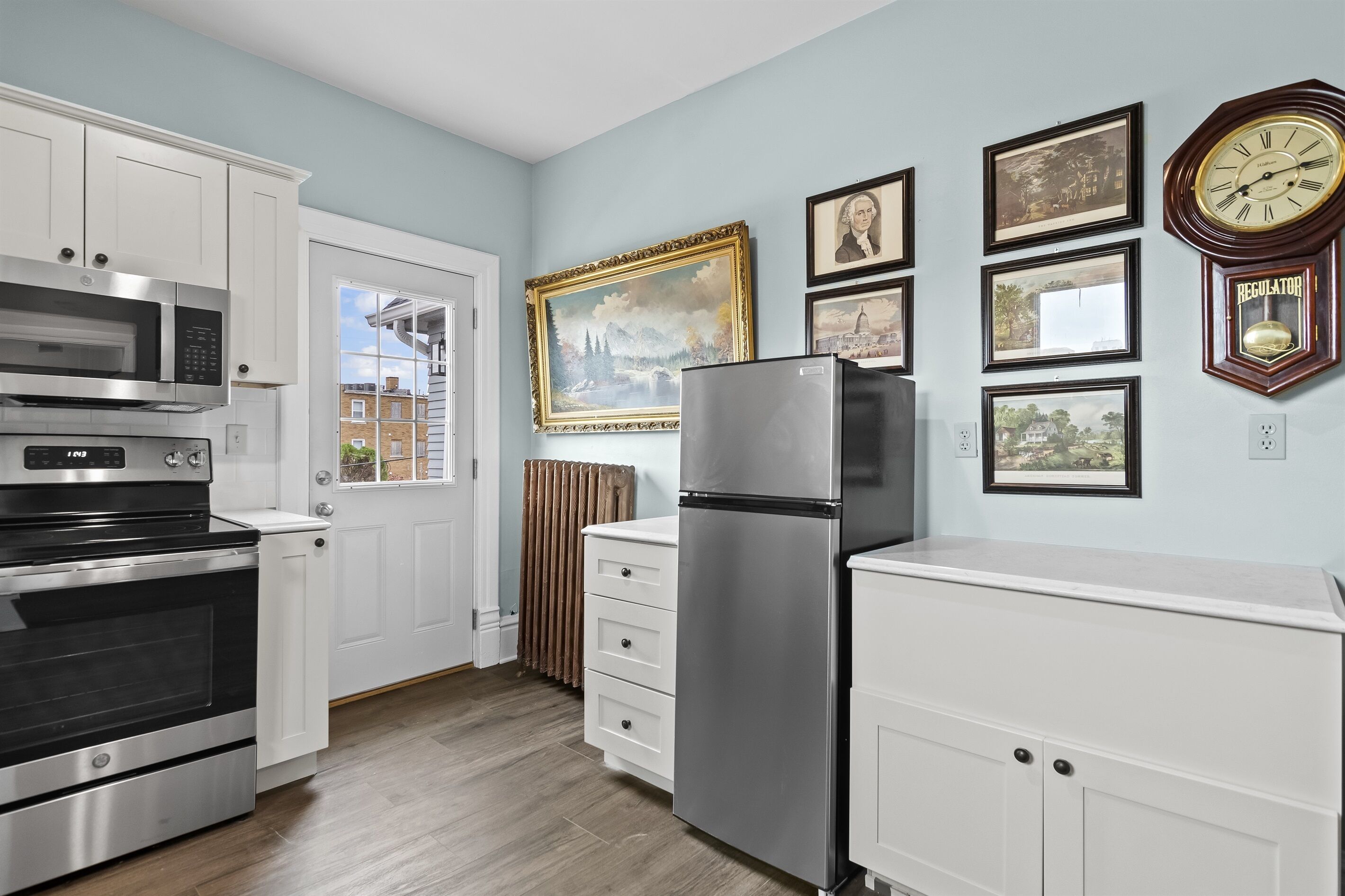 A kitchen with white cabinets, stainless steel appliances, and a refrigerator. There are framed pictures on the wall and a clock.