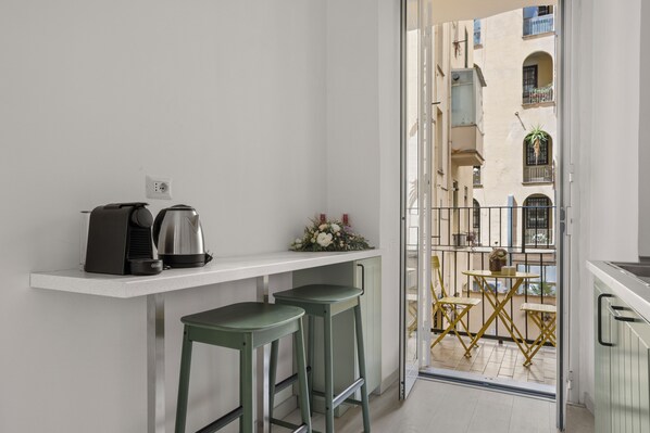 A small kitchen with a coffee maker, two stools, and a balcony with yellow chairs and a table.