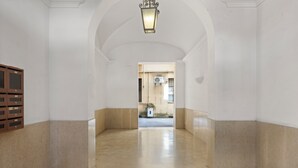 A hallway with white walls and beige flooring, leading to an open area with a potted plant and an air conditioning unit.