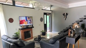 A modern living room with a flat-screen TV, black leather sofas, a fireplace, and a staircase. - Casa de Praia com Trilha Privativa em Condomínio Fechado (Florianópolis)