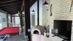 A pool table, a brick oven pizza oven, and a fireplace in a room with brick walls and wooden beams. - Casa de Praia com Trilha Privativa em Condomínio Fechado (Florianópolis)