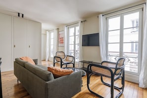A modern living room with a grey sofa, two black wicker chairs, a small table, and a TV mounted on the wall. - Bright & Stylish Two-Bedroom Apartment with Modern Comforts in Saint-Germain (Paris)