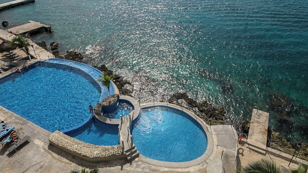 A large, circular swimming pool with a clear view of the sea, a small adjacent pool, and a stone deck with lounge chairs.