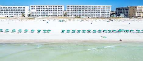 A beachfront hotel with multiple floors, a sandy beach with umbrellas, and a clear view of the ocean.