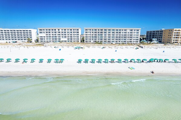 A beachfront hotel with multiple floors, a sandy beach with umbrellas, and a clear view of the ocean.
