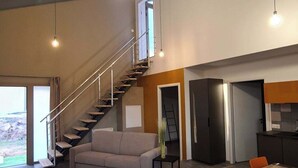 A spacious living area with a high ceiling, a staircase leading to an upper level, a large dining table with chairs, and a kitchen area.