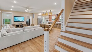 A spacious living room with a large sectional sofa, a flat-screen TV, and a dining area with a kitchen in the background. - Creamsicle Cottage: Newly Built | Saltwater Pool (Corolla)