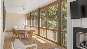 A covered outdoor area with a wooden deck, a round table with chairs, a TV mounted on the wall, and a fireplace. - Creamsicle Cottage: Newly Built | Saltwater Pool (Corolla)