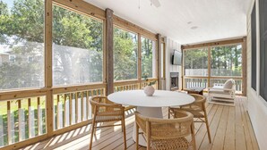 A covered outdoor patio with a round table and wicker chairs, large windows, and a ceiling fan. - Creamsicle Cottage: Newly Built | Saltwater Pool (Corolla)