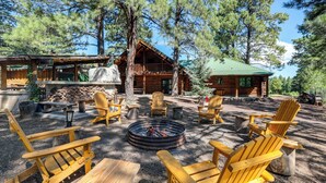 A rustic cabin with a fire pit surrounded by Adirondack chairs, a wooden deck, and a large tree in the center.