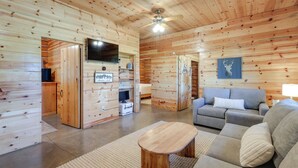 A log cabin interior with a sofa, a wooden coffee table, a ceiling fan, and a mounted television.
