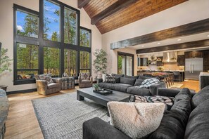 A spacious living area with a large sofa, a coffee table, and a fireplace. There are multiple windows providing a view of the outdoors. - Forest View Hideaway with Access to Forest House (Sunriver)