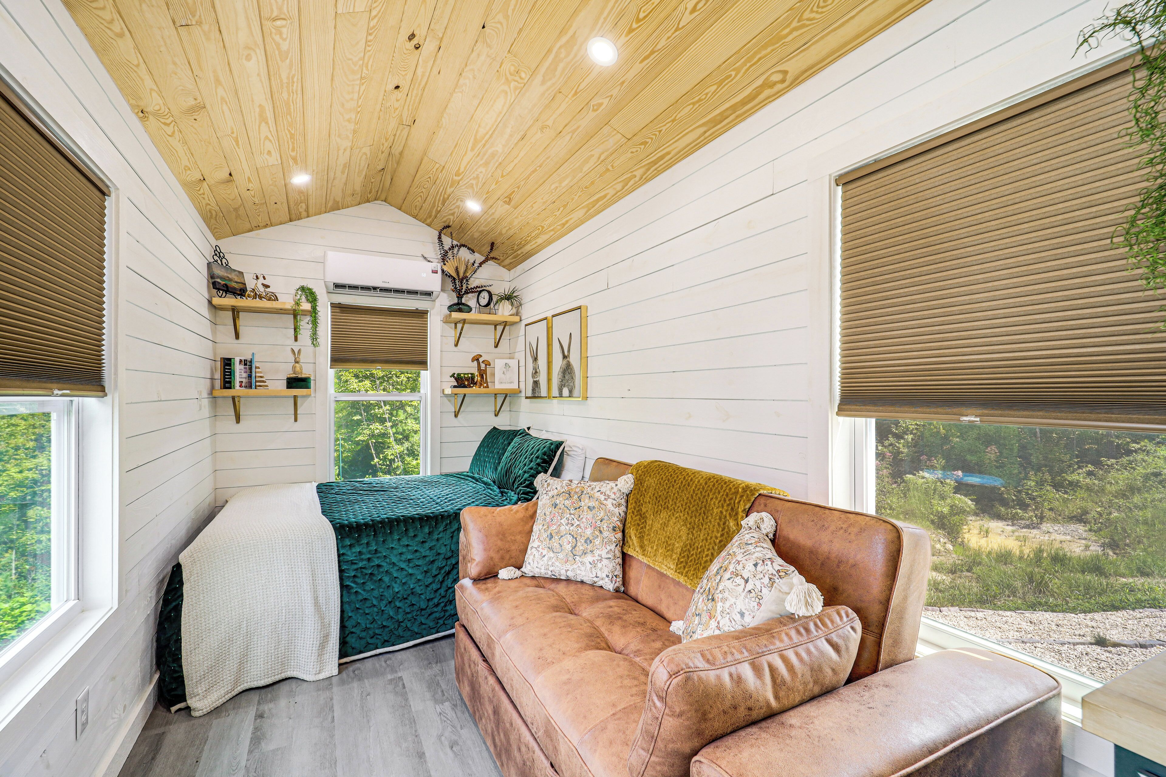 A cozy room with a wooden ceiling, a brown leather sofa, a green patterned bed, and a window with a view of greenery.