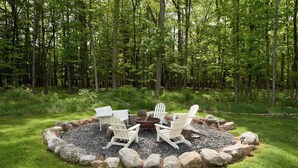 Ein kreisförmiger Sitzbereich mit Adirondack-Stühlen und einer Feuerstelle, umgeben von Steinen, in einer grünen Wiese mit einem Wald im Hintergrund.
